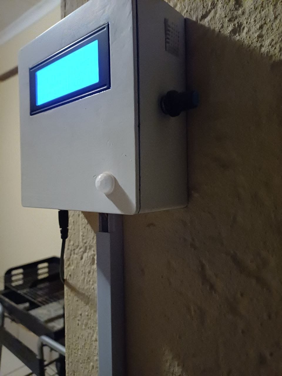 The pool monitor mounted on the wall in its painted white 3D printed enclosure, with the blue LCD backlight lit, the PIR sensor dome below it and the power lead dropping away underneath