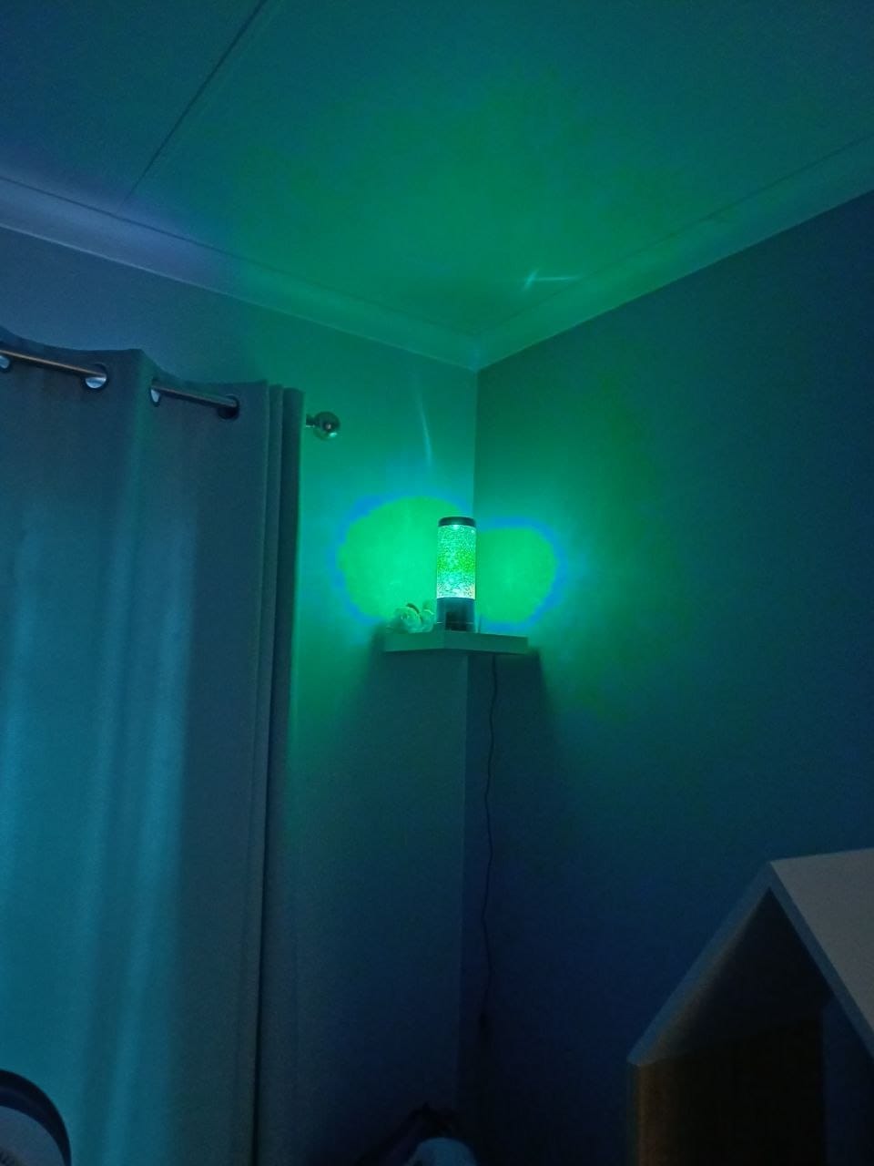 The glitter lamp glowing green on the corner shelf above her bed, lighting the wall and ceiling green