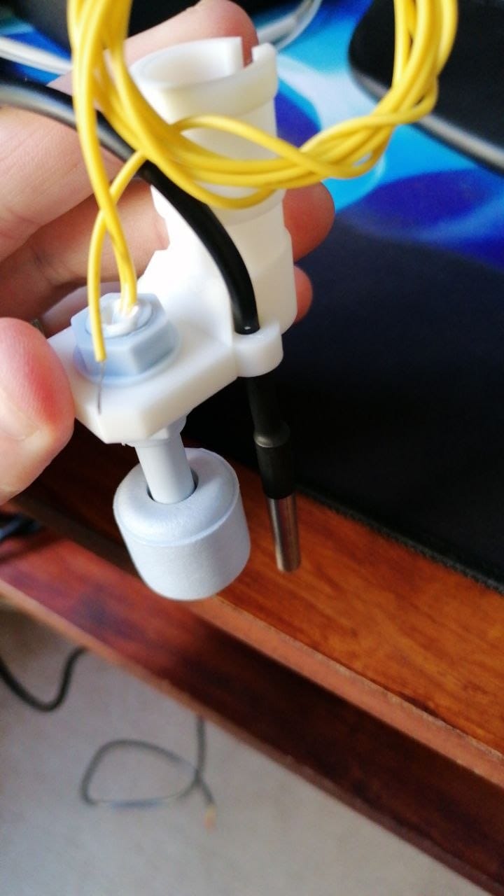 Holding the assembled bracket, with the float switch hanging below it and the black temperature probe cable beside it