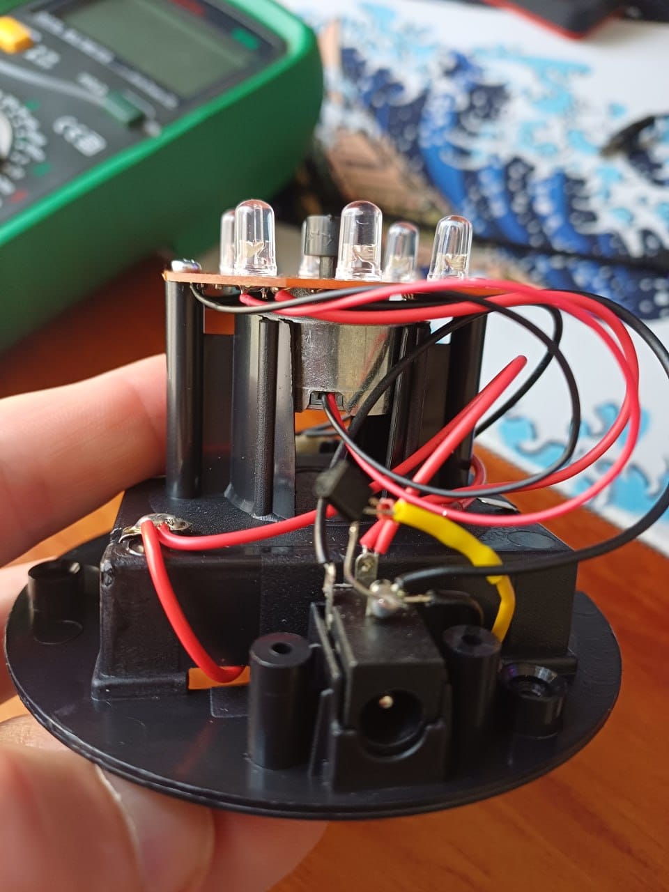 Lamp internals with wiring, motor, power jack, a transistor and a multimeter in the background