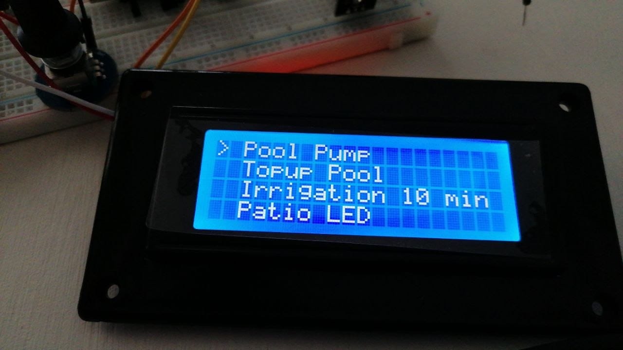 The custom menu on the 20x4 LCD reading Pool Pump, Topup Pool, Irrigation 10 min and Patio LED, with the selection arrow on the first item, on the bench with the breadboard and rotary encoder behind it