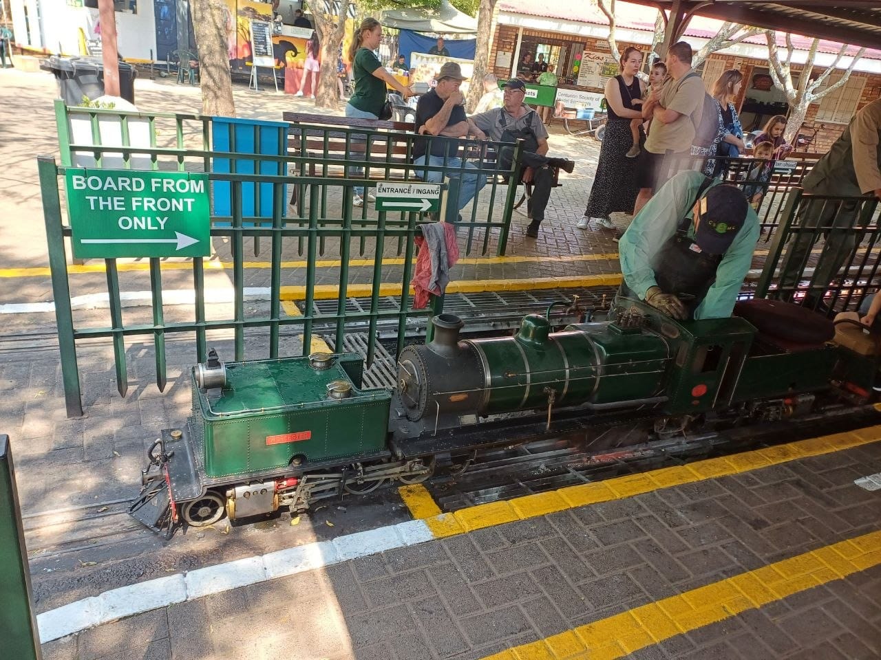 A green ride-on steam locomotive at the platform with a driver in green overalls leaning over the cab, passengers waiting behind the fence
