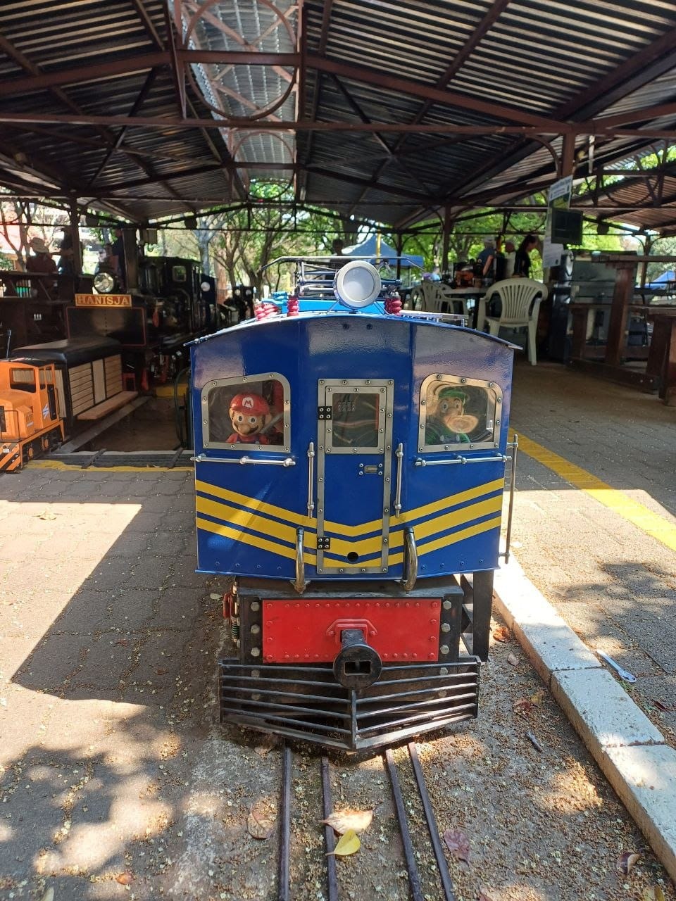The front of a blue ride-on locomotive with yellow chevrons and a red buffer beam, with two plush toys looking out of the cab windows