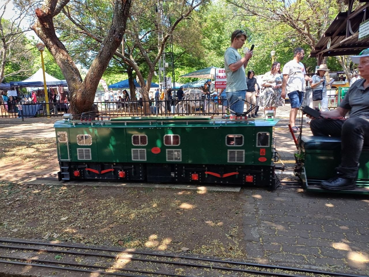 A green ride-on electric locomotive in the yard lettered C.S.M.E., with people watching from the platform