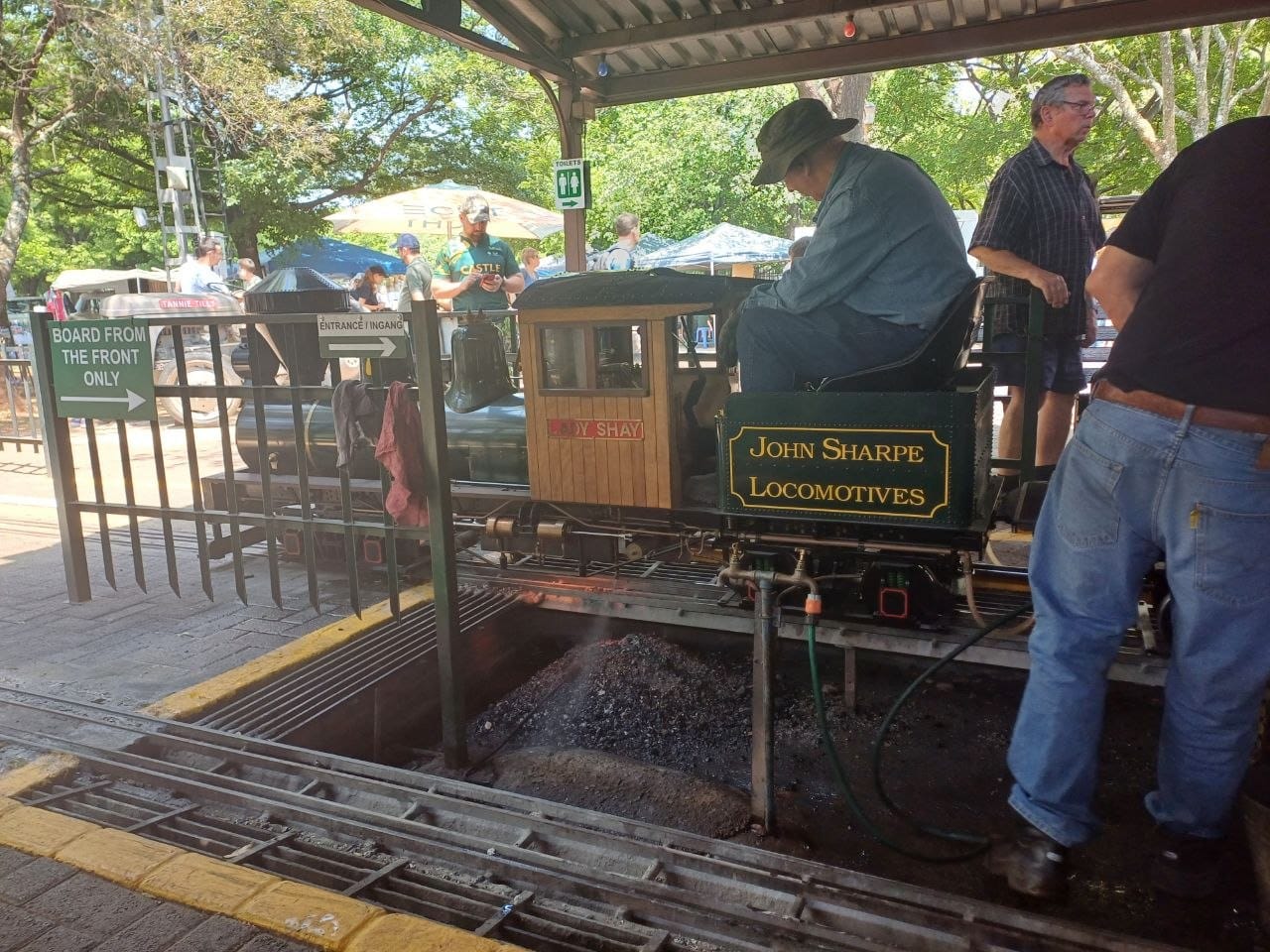 A ride-on steam locomotive at the platform with a driver in overalls and a wide hat, passengers waiting under the shade structure