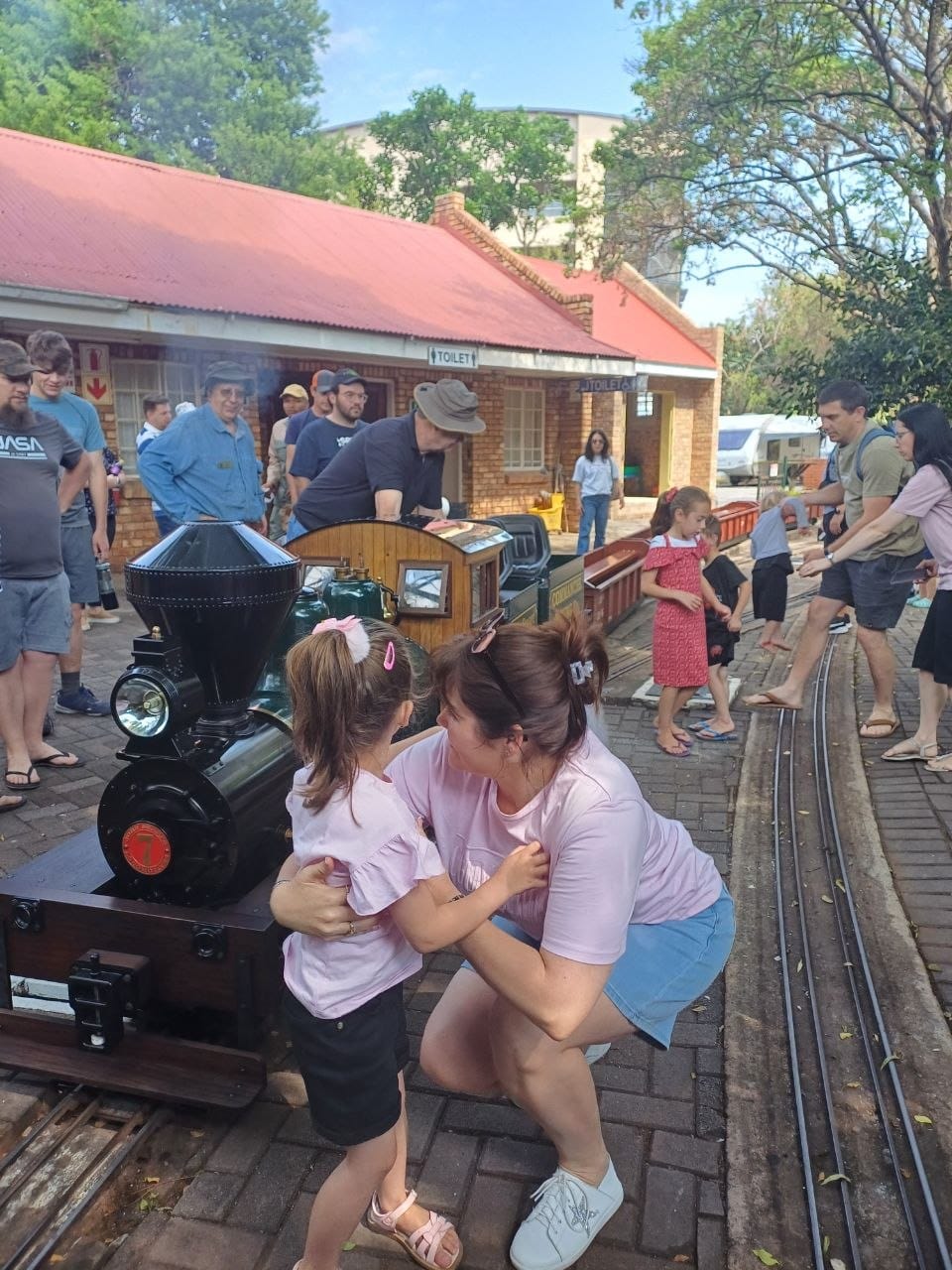 Cheryl crouching down to hug Ava beside the black steam locomotive at the station platform, with the station building and its red roof behind