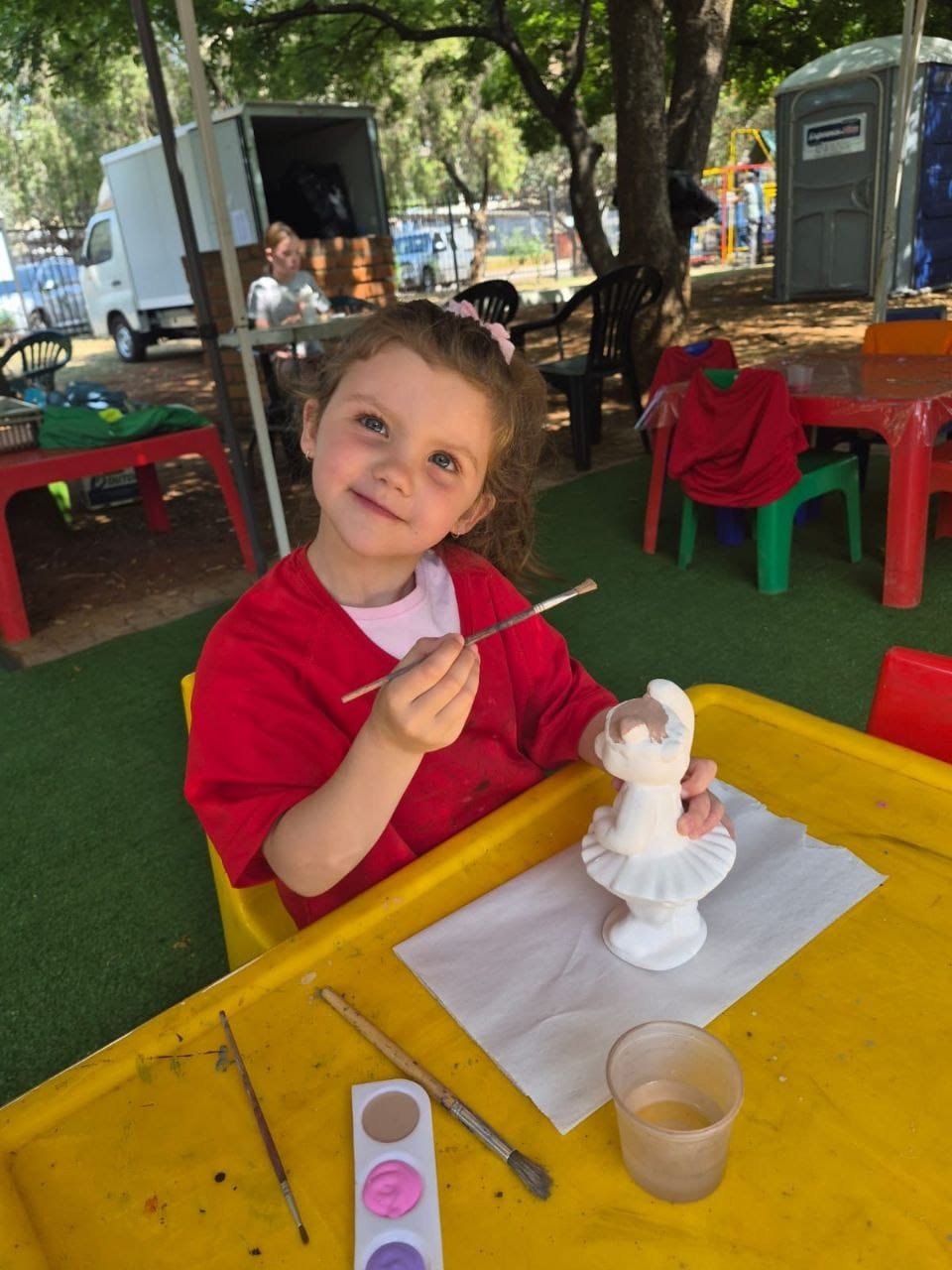 Ava at the porcelain painting table in a red smock, painting a white ballerina figurine