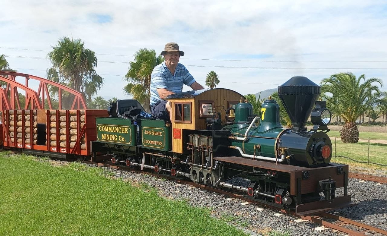 A dark green ride-on steam locomotive lettered Commache Mining Co and John Sharpe Locomotives, with a driver in a sun hat, crossing a red steel bridge with a timber wagon behind