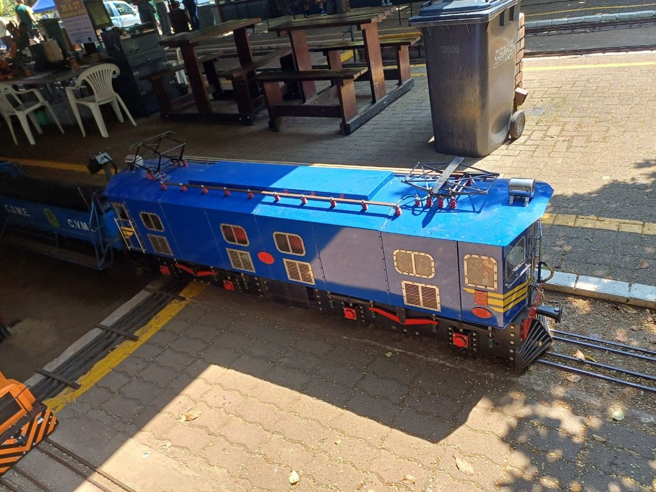 A blue ride-on electric locomotive lettered C.S.M.E. on the track beside the picnic benches