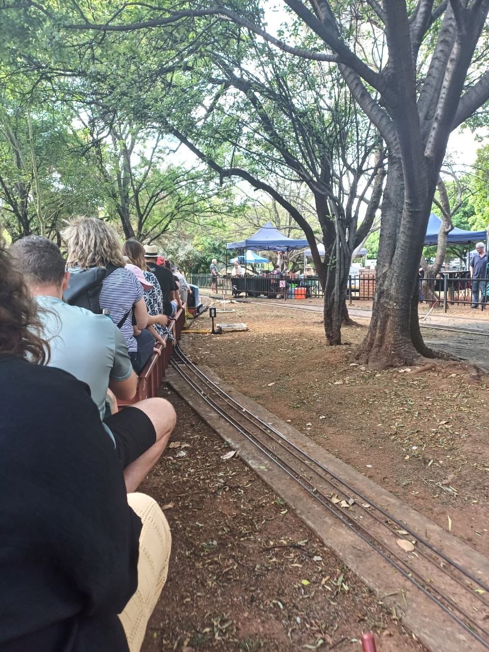 Looking along the train from behind, rows of passengers in the open carriages, narrow gauge track on the ground beside the train and blue gazebos in the distance
