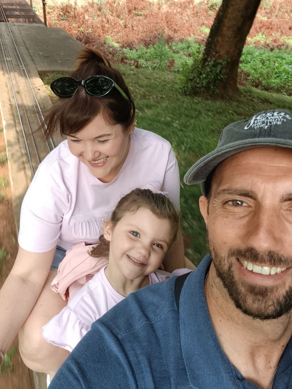 Selfie on the moving train with Collin in a cap and blue polo in front and Cheryl and Ava behind, trees blurring past and the track curving away