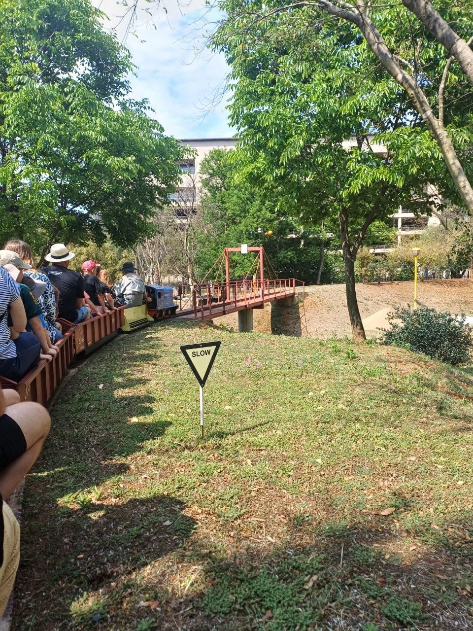 The train crossing the red suspension bridge with a large SLOW sign in the grass and passengers in the open carriages