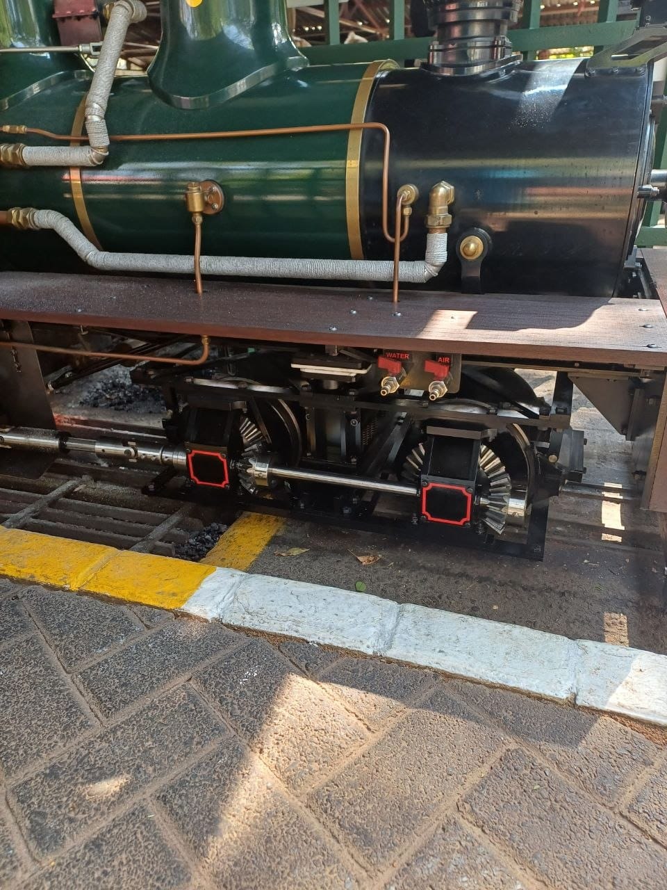 Close-up of a ride-on steam locomotive's green boiler, brass fittings and valve gear above the yellow platform edge