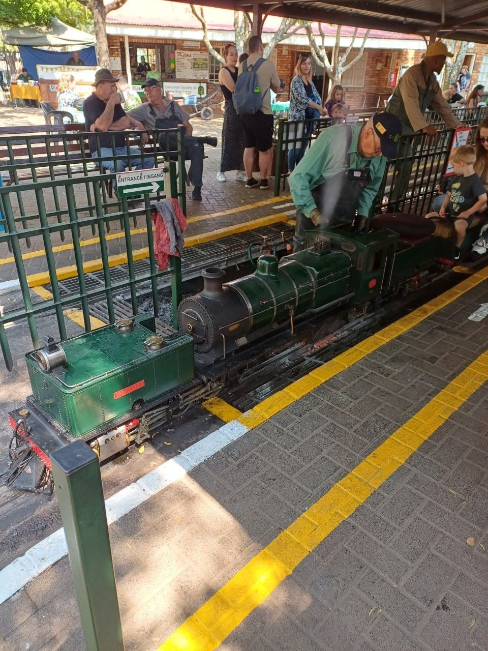 A green ride-on steam locomotive being driven along the platform with a young passenger in the carriage behind