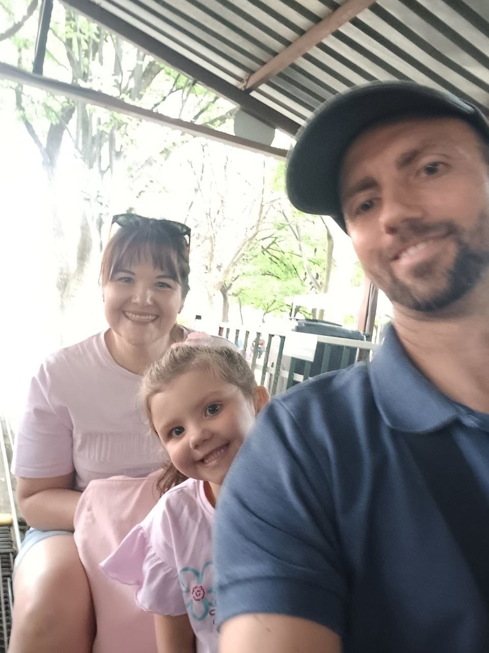Selfie of the three of them under the station canopy with the corrugated roof overhead and trees beyond