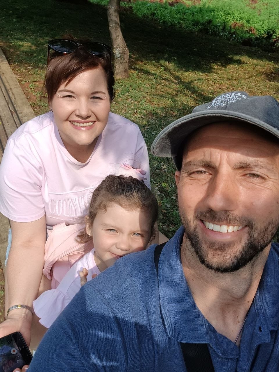 Family selfie on the grass with Collin in a cap and blue polo, Cheryl in pink, and Ava between them