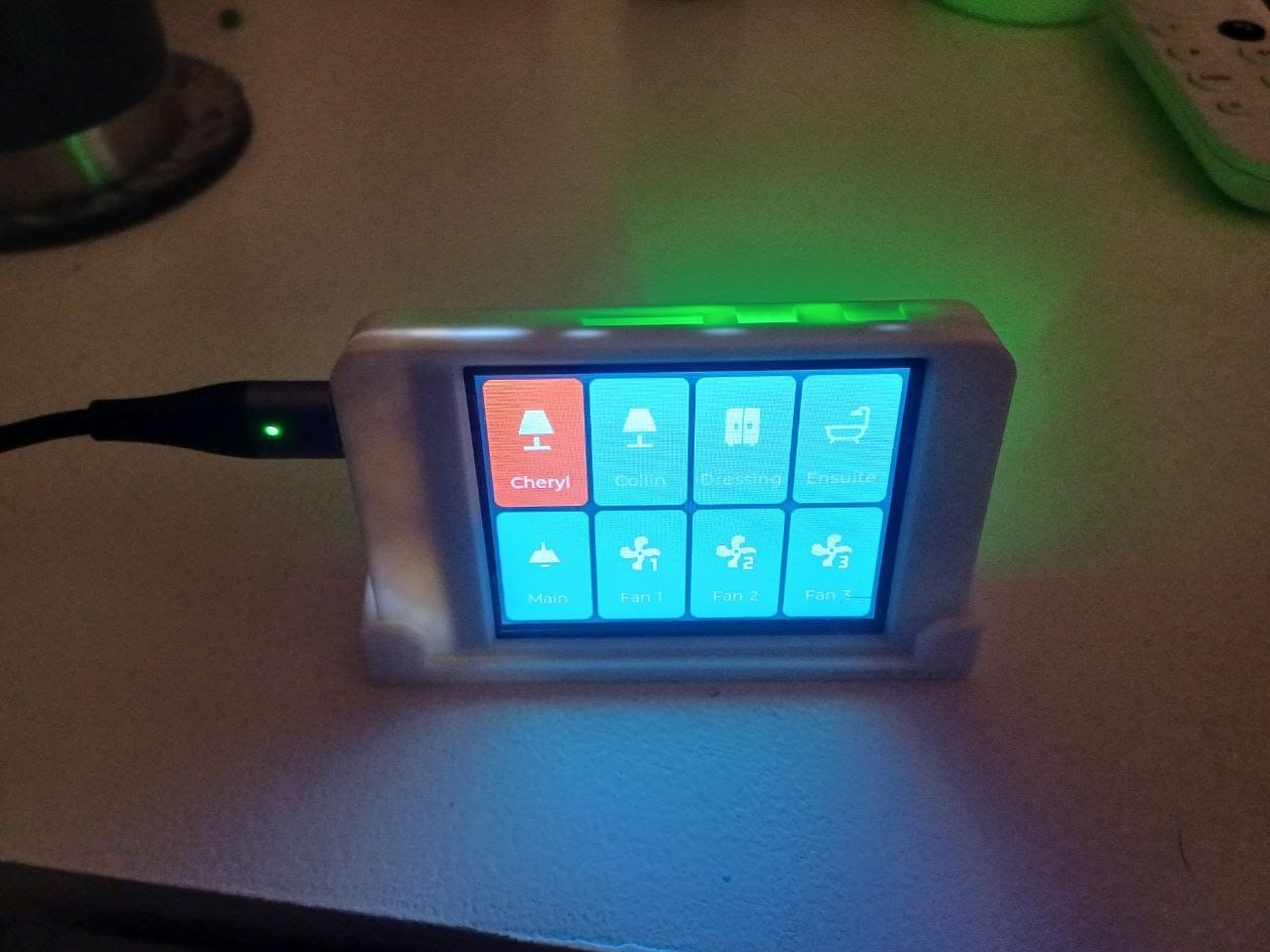 A small ESP32 touch display in a white 3D-printed case on a bedside table, showing eight tiles labelled Cheryl, Collin, Dressing, Ensuite, Main, Fan 1, Fan 2 and Fan 3, with the Cheryl tile lit red and a green glow spilling from the case